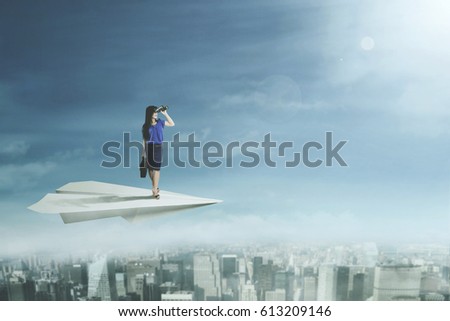 Picture of a young businesswoman looking through binoculars while flying with a paper plane above a city