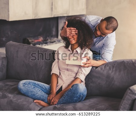 Beautiful young Afro American couple on couch at home. Handsome guy is covering his girlfriend eyes and giving a gift box