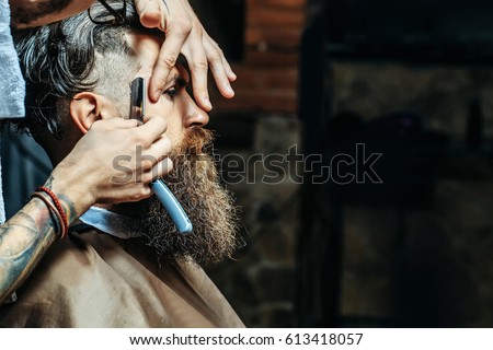 Bearded man with long beard, brutal, caucasian hipster with moustache, getting stylish hair shaving, haircut, with razor by barber or hairdresser with tattoo on hands at barbershop Royalty-Free Stock Photo #613418057