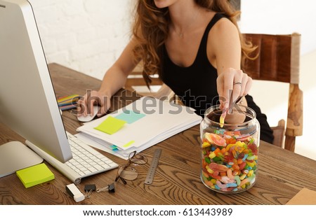 Cropped image of young stressed woman eating sweets at workplace in office. The girl takes candy from big glass jar with lollipops standing on a desktop. Stress and junk food concept Royalty-Free Stock Photo #613443989