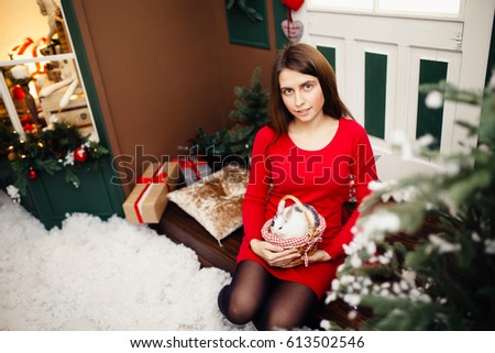 European girl in red dress hold in arms little white rabbit with grey area against the backdrop of New Year's tree.