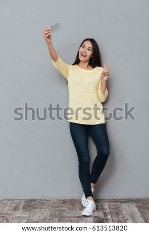 Smiling cute young woman taking selfie with smartphone and showing peace sign over grey background