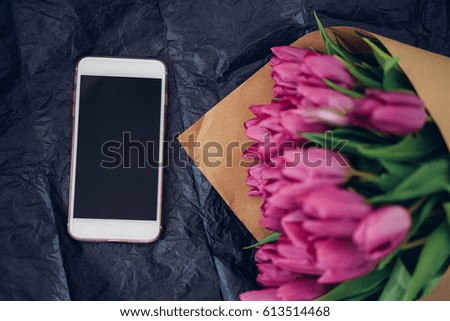 Bouquet of purple tulips in craft paper and white smart phone on the gray wooden background. Top view. Copy space. Presentation template. 