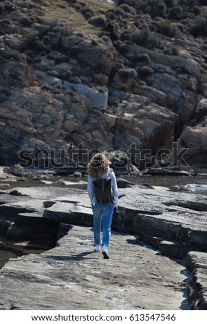 Full length portrait of blond woman tourist with khaki backpack wearing jeans and gray jacket walking on rock ribbed shore