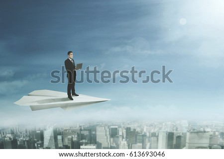 Picture of a young businessman standing on a big paper plane while using a laptop computer and flying above a city 