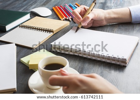 Close up of lady's hands writing in empty notepad placed on office desktop with other supplies and coffee cup. Copy space
