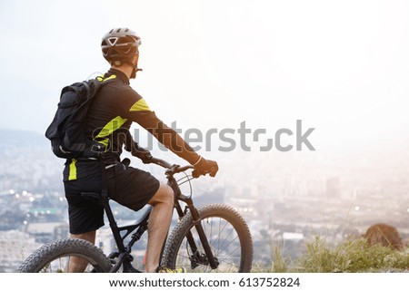 Rear shot of young rider standing on top of hill with booster bike and looking down at foggy city while biking in the morning. Cyclist in black and yellow sports clothes admiring beautiful view