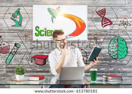 Attractive caucasian male reading book at workplace with creative sketch, laptop, supplies and other items. Knowledge concept 