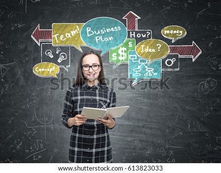 Portrait of a smiling nerdy girl wearing glasses and a black checkered shirt and holding a book while standing near a blackboard with a business plan scheme.