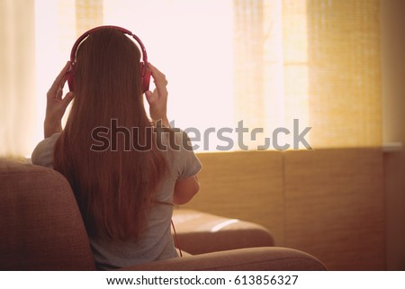 Happy woman with pink headphones listening to music. Young woman sit backwards at home on a couch and enjoy the music with headphones near sunny window