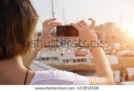 Young woman take a photo of a sea port in a rays of sunset. Focus on female hands with smart phone. Flare light.                               