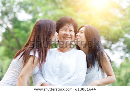 Portrait of Asian daughters kissing elderly mother, senior adult woman and grown child. Outdoors family at nature park with beautiful sun flare.