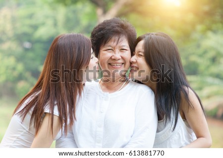 Portrait of beautiful Asian daughters kissing elderly mother, senior adult woman and grown child. Outdoors family at nature park with beautiful sun flare.