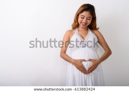 Studio shot of young happy Asian pregnant woman smiling while making hand heart sign on her stomach with love for her baby against white background