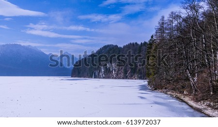 Frozen Lake Alpsee in the alps in Bavaria, Germany. Taken in winter, showing the surrounding pine forests and high mountains on a sunny day.