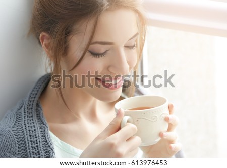 Beautiful young woman drinking tea near window at home Royalty-Free Stock Photo #613976453