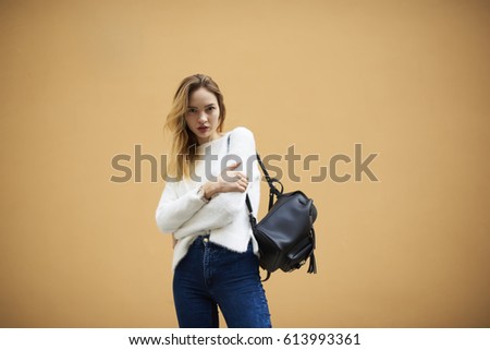 Half length portrait of good looking beautiful model with blond hair looking at camera. Hipster girl in street wear with style backpack on shoulder posing near promotional background for advertise