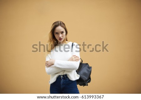 Half length portrait of attractive fashionable female dressed in white sweatshirt with black backpack on shoulders serious looking on camera and posing against promotional background for advertise