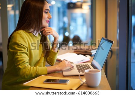 Charming young hipster girl in good mood chatting in social networks with friends while working on freelance creating graphic design for websites using laptop computer and free wireless connection