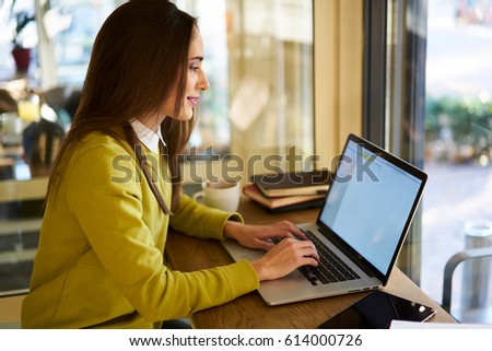 Smiling attractive good looking female journalist working on freelance monitoring news on online issue web site sitting in coffee shop using laptop computer and free wireless connection to internet