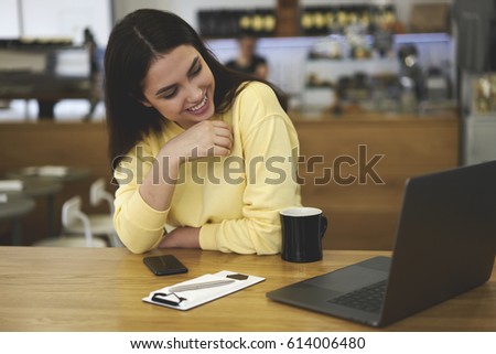 Young cheerful hipster girl laughing after looking funny videos on modern laptop received from best friends in social networks sitting in designer cafeteria interior enjoying cup of aroma coffee