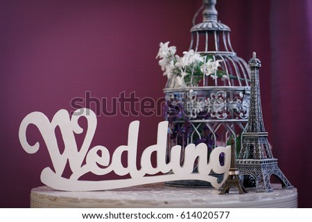 The word "Wedding" made of white wooden letters, statuettes of the Eiffel Tower, and cage for birds, stand on a table on a purple background