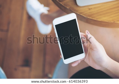 Mockup image of hands holding white mobile phone with blank black screen on thigh with silver colour laptop on table and wooden floor background