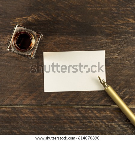 An overhead photo of a blank business card on a dark wooden background texture with a golden ink pen and ink bottle. A square mockup or a banner with copy space
