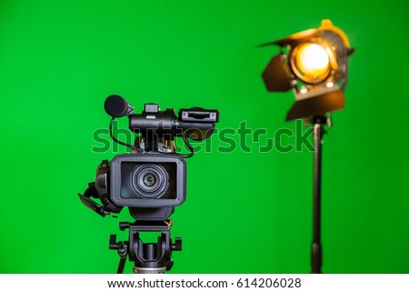 A video camera and a spotlight with a Fresnel lens on a green background. Filming in the interior. The chroma key.
