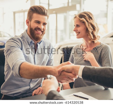 Visiting car dealership. Beautiful couple is talking to sales manager and smiling. Men are shaking hands