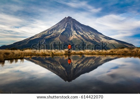 A mountaineer with an orange dress in a symmetric picture looking to the taranaki volcano in the north island of new zealand and with the reflection of tho mountain and the climber in a lake. Zeland Royalty-Free Stock Photo #614422070