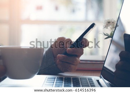 Casual man using smart phone and laptop computer, web browsing internet at home and drinking coffee in the morning light with reflection on laptop computer screen, internet browsing on phone concept