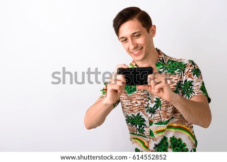 Studio shot of young happy tourist man smiling while taking picture with mobile phone against white background