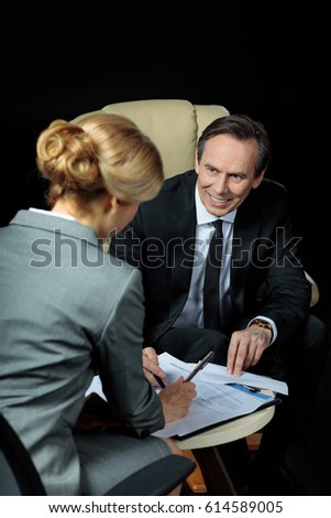 Smiling mature businessman looking at blonde businesswoman signing papers  isolated on black