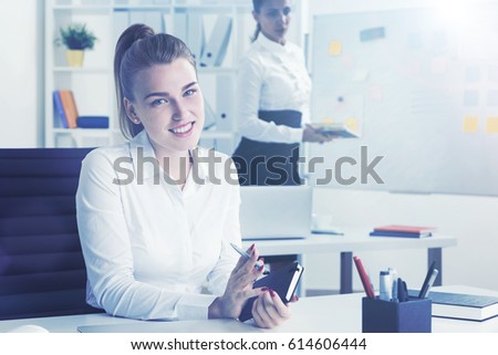 Portrait of a smiling blond businesswoman with notebook at her workplace in a white office. Her colleague is in the background making a drawing on a whiteboard. Toned image