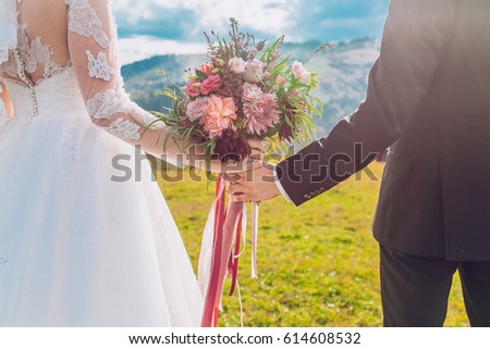 Closeup view of married couple holding hands and carrying bride's bouquet behind mountains and nature landscape. Advertising picture. Copy space. Amazing colors.