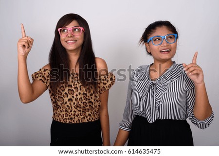 Studio shot of two happy young Persian woman friends smiling while thinking and pointing finger up with eyeglasses together against gray background