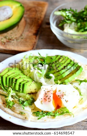 Poached egg, avocado slices, salad mix, sauce, spices in a flour tortilla. Avocado half on a wooden board, salad mix in a glass bowl on a table. Healthy and diet breakfast idea. Vertical photo