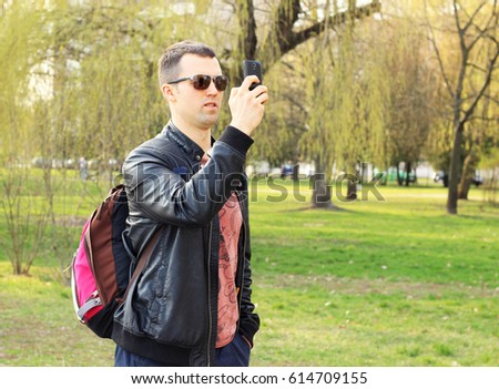man in sunglasses with a backpack as a tourist photographs in surprise on the black phone in a city green park