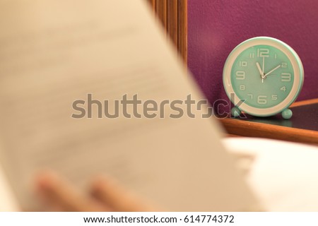 Image of teal alarm clock at 11.00 p.m. night time with blurry hand holding bible book as woman reading textbook at night on white bed in bedroom.World Book Night in April 23rd.