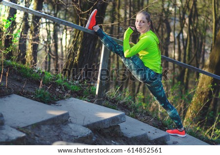 Fit teen girl athlete doing leg split stretching exercises outdoors in a park.