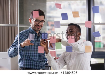 Two young multiethnic businessmans are working in office on glass wall with sticky notes
