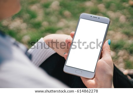 Girl with smartphone in hands with mock up white screen of blank screen sits in park on open space, screen for content integration. Hands holding gadget on blurred backdrop, front view