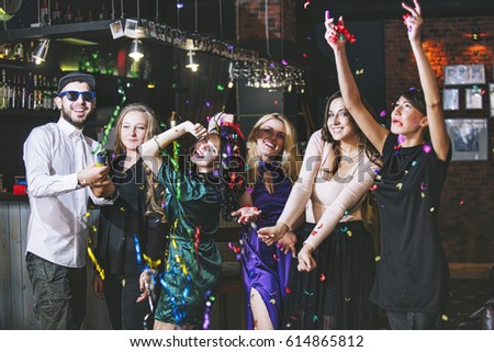 Young cheerful company of friends in the club bar having fun with multi-colored confetti and crackers celebrate the holidays