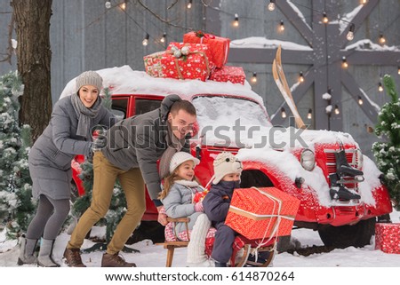 A beautiful family (mother, father, daughter and son) ride a sleigh, a snowy day. On the background of a red car. Gifts and Christmas trees.