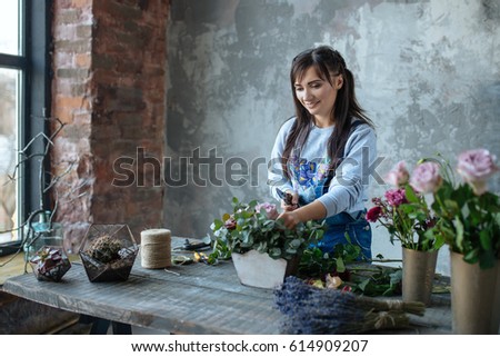 female  in gray blouse and jeans make a bouquet over gray background, putting roses in vase, flowers and vase on wood table, workplace