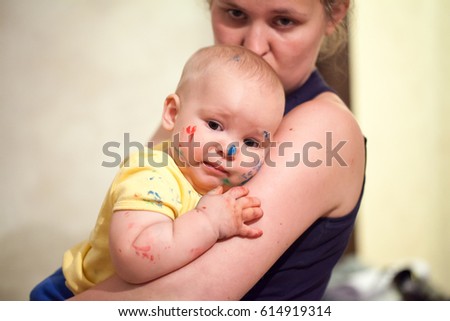 The portrait of a little dirty baby, a boy, painted with paints, sadly sits on the hands of a young woman, mother