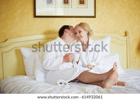 Happy young couple in white bathrobes drinking coffee together in bed at morning. Hotel, travel, relationships, and happiness concept