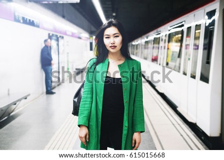 Young beautiful stylish asian lady with long dark hair wearing long green jacket is looking at the camera while standing on a blurred metro platform background while train is coming.