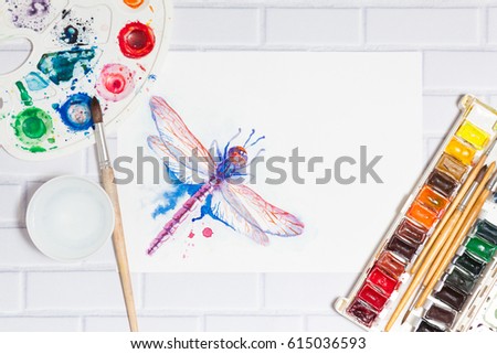 Hand Drawn Bright Sketch of Violet Dragonfly, with lying paints, paintbrushes and palette on the white brick background - concept of human creativity, top flat view with place for text